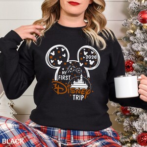 May include: Black sweatshirt with a white and brown Mickey Mouse design. The design includes the text "MY FIRST DISNEY TRIP" and the year "2026". The sweatshirt is paired with plaid pants and a white mug.