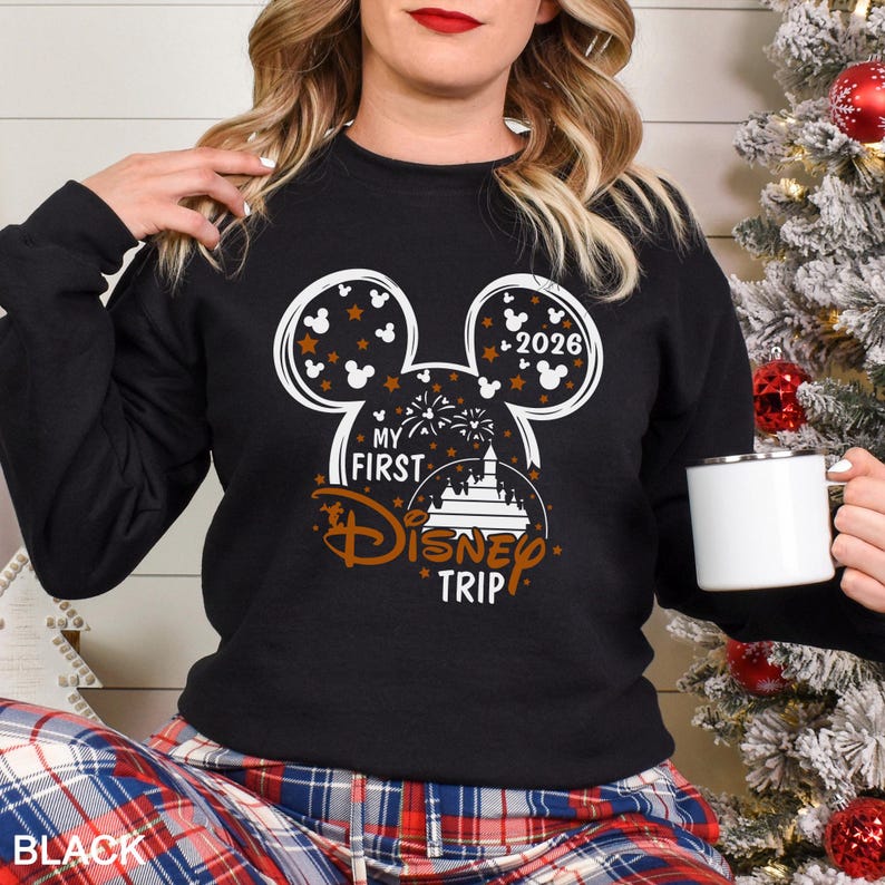 May include: Black sweatshirt with a white and brown Mickey Mouse design. The design includes the text "MY FIRST DISNEY TRIP" and the year "2026". The sweatshirt is paired with plaid pants and a white mug.