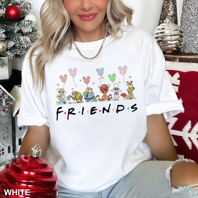 K&ouml;nnte beinhalten: Wei&szlig;es T-Shirt mit einem farbenfrohen Aufdruck von Zeichentrickfiguren, die Mickey Mouse-f&ouml;rmige Ballons &uuml;ber dem Wort "FRIENDS" in Schwarz halten. Das Design umfasst verschiedene Charaktere und Sterne. Das Shirt ist ein l&auml;ssiger Unisex-Stil.