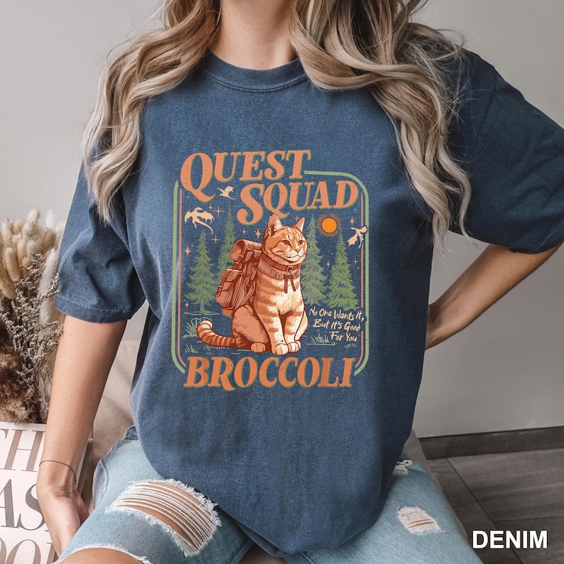 May include: Denim blue t-shirt featuring a graphic of an orange tabby cat with a backpack, set against a forest scene. The text reads "QUEST SQUAD BROCCOLI" with the phrase "No One Wants It, But It's Good For You."