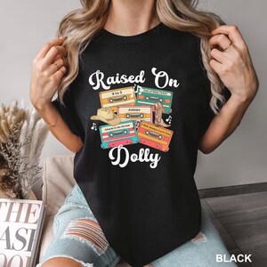 May include: A black t-shirt featuring a graphic print with the text "Raised On Dolly" above and below four colorful cassette tapes labeled with song titles, a cowboy hat, and a cowboy boot, all arranged in a central cluster on the chest.