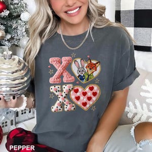 May include: A charcoal gray short-sleeve t-shirt featuring a graphic print of two cartoon characters inside a heart, alongside the letters X and O in a textured knit pattern. The design includes small red hearts, gold stars, and polka dots.