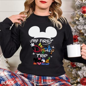 Puede incluir: Sudadera negra con un dise&ntilde;o de cabeza de Mickey Mouse blanca y el texto "MY FIRST DISNEY TRIP 2025" en letras coloridas. La persona lleva la sudadera y sostiene una taza blanca.