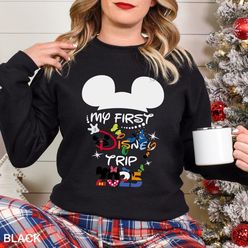 Puede incluir: Sudadera negra con un dise&ntilde;o de cabeza de Mickey Mouse blanca y el texto "MY FIRST DISNEY TRIP 2025" en letras coloridas. La persona lleva la sudadera y sostiene una taza blanca.