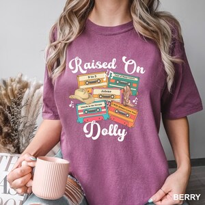 May include: A berry-colored short-sleeve t-shirt featuring the white text "Raised On Dolly" and a graphic of four colorful cassette tapes labeled "9 to 5," "Jolene," "Coat of Many Colors," and "I Will Always Love You," with a cowboy hat and boot.