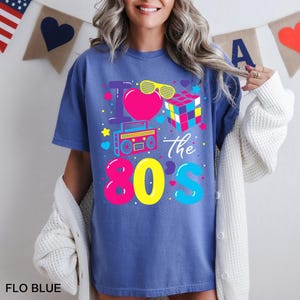 May include: A blue t-shirt featuring a graphic print with the text I Love the 80s. The design includes a pink heart, yellow sunglasses, a colorful Rubik's cube, and a cyan boombox, surrounded by small stars, hearts, and dots in bright colors.