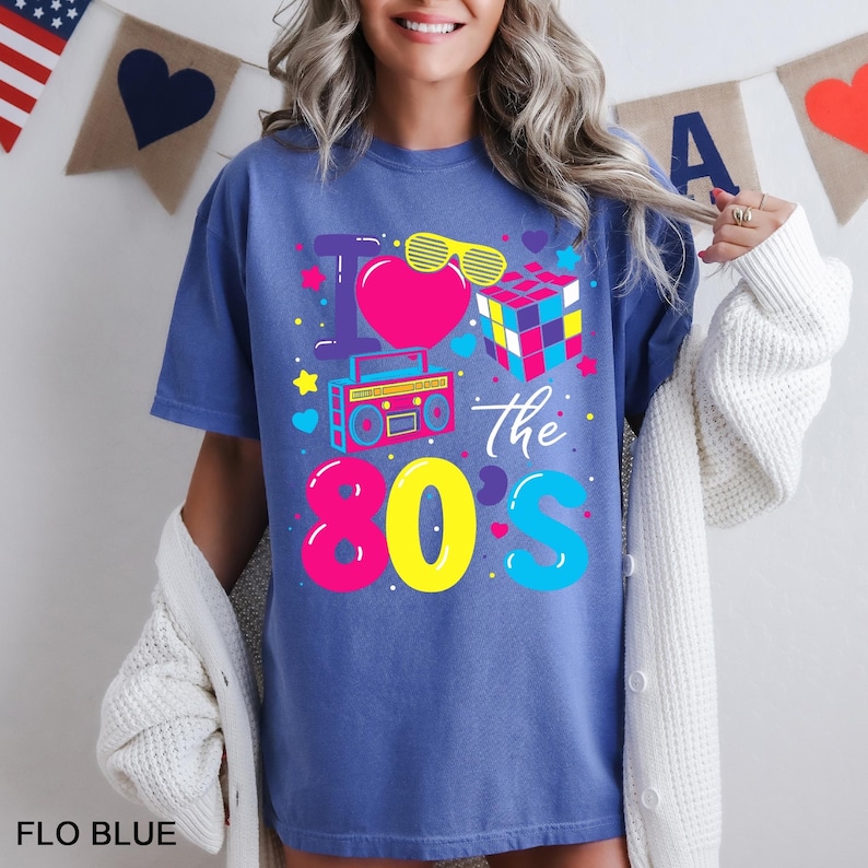 May include: A blue t-shirt featuring a graphic print with the text I Love the 80s. The design includes a pink heart, yellow sunglasses, a colorful Rubik's cube, and a cyan boombox, surrounded by small stars, hearts, and dots in bright colors.
