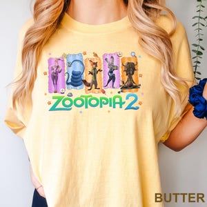 K&ouml;nnte beinhalten: Hellgelbes T-Shirt mit einem farbenfrohen Aufdruck von Charakteren aus "Zootopia 2". Das Design zeigt einen Geparden, eine Schlange, einen Fuchs, ein Kaninchen und einen Yak. Der Text "Zootopia 2" ist in Gr&uuml;n und Blau gedruckt.