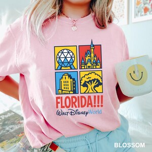 Może przedstawiać: Jasnor&oacute;żowa koszulka z kolorową grafiką przedstawiającą zabytki Florydy, w tym kulę, zamek, budynek i drzewo. Tekst "FLORIDA!!! Walt Disney World" jest wydrukowany poniżej. W dłoni trzymany jest biały kubek z ż&oacute;łtą buźką.