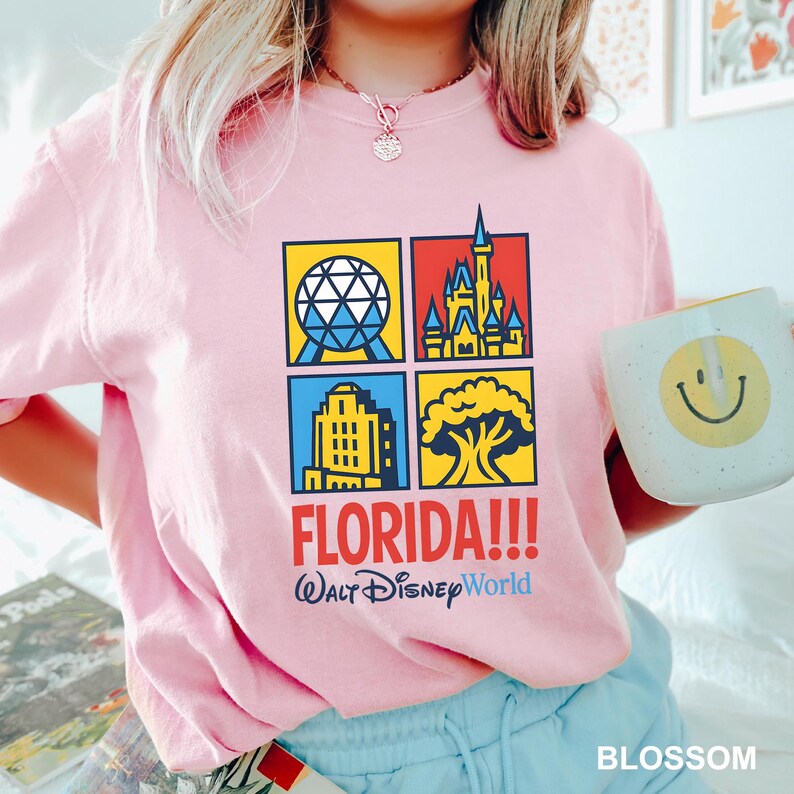 Może przedstawiać: Jasnor&oacute;żowa koszulka z kolorową grafiką przedstawiającą zabytki Florydy, w tym kulę, zamek, budynek i drzewo. Tekst "FLORIDA!!! Walt Disney World" jest wydrukowany poniżej. W dłoni trzymany jest biały kubek z ż&oacute;łtą buźką.