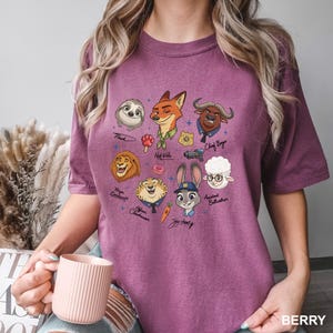 K&ouml;nnte beinhalten: Beerenfarbenes T-Shirt mit Zeichentrickfiguren aus Zootopia. Das Design umfasst Flash, Nick Wilde, Chief Bogo, Mayor Lionheart, Officer Clawhauser, Judy Hopps und andere. Das Wort "BERRY" ist unten rechts auf dem Shirt aufgedruckt.