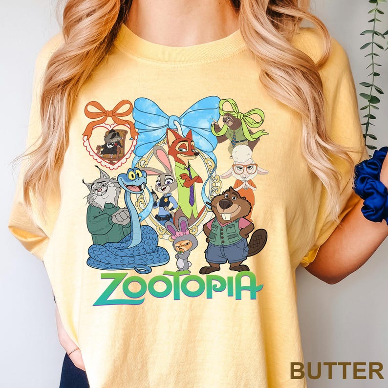 K&ouml;nnte beinhalten: Hellgelbes T-Shirt mit einem farbenfrohen Aufdruck von Charakteren aus dem Film "Zootopia". Das Design umfasst eine blaue Schleife, einen goldenen Rahmen und das Wort "Zootopia" in Gr&uuml;n und Blau. Das T-Shirt hat eine hellgelbe Farbe.