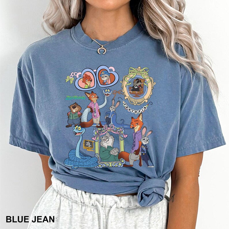 May include: A blue crew neck t-shirt featuring a graphic print of Zootopia characters including Nick Wilde, Judy Hopps, and Flash. The design includes various framed portraits, heart-shaped outlines, and the text Zootopia in a colorful font.