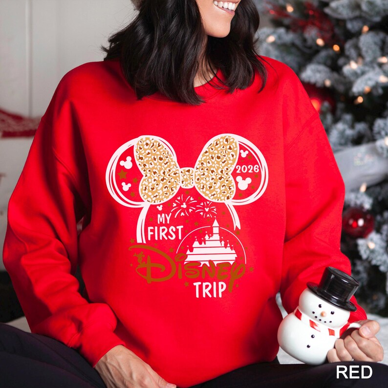 May include: A red sweatshirt with a white and gold graphic design. The design includes Minnie Mouse ears, a bow with a leopard print pattern, the text "MY FIRST DISNEY TRIP", and the year "2026".