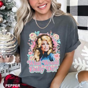 May include: A charcoal gray short-sleeve t-shirt featuring a graphic print of a blonde woman surrounded by pink and yellow flowers and blue butterflies. The text "What Would Dolly Do?" is printed in a pink and white retro-style font below the image.