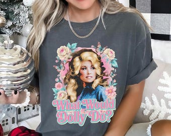 Può includere: Una t-shirt grigio antracite a maniche corte con una stampa grafica di una donna bionda circondata da fiori rosa e gialli e farfalle blu. Il testo "What Would Dolly Do?" è stampato in un carattere retro rosa e bianco sotto l'immagine.