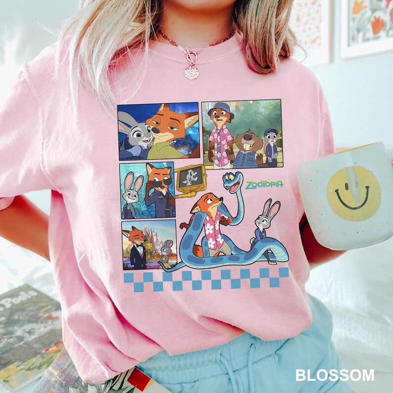 May include: A light pink t-shirt featuring a graphic print of Zootopia characters including Judy Hopps and Nick Wilde in various scenes. The design includes a blue snake, a checkered pattern at the bottom, and the Zootopia logo in green text.