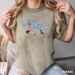 Op de afbeelding: Khaki T-shirt met een cartoon illustratie van een vos en een konijn die rennen met een blauwe slang. De vos draagt een roze shirt en een bruine broek, terwijl het konijn een blauw pak draagt. Het woord "KHAKI" staat onderaan gedrukt.