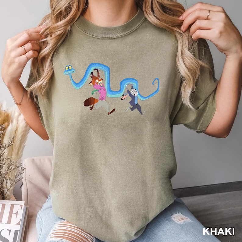 Op de afbeelding: Khaki T-shirt met een cartoon illustratie van een vos en een konijn die rennen met een blauwe slang. De vos draagt een roze shirt en een bruine broek, terwijl het konijn een blauw pak draagt. Het woord "KHAKI" staat onderaan gedrukt.