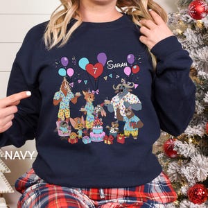 May include: Navy blue sweatshirt featuring a colorful cartoon illustration of Zootopia characters celebrating a birthday. The design includes balloons, a cake, and the name "Sarah" with the number "7". The word "NAVY" is printed on the bottom left.