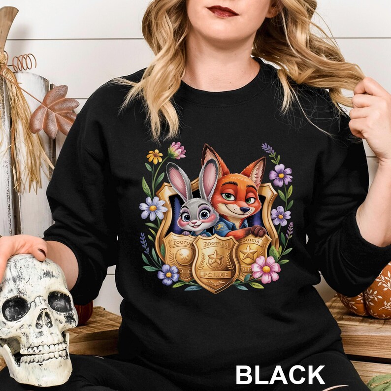 May include: Black crewneck sweatshirt featuring a colorful graphic of Judy Hopps and Nick Wilde from Zootopia, framed by flowers and police badges. The word "BLACK" is printed at the bottom. A fun, animated design.