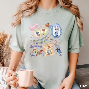 Op de afbeelding: Lichtgroen T-shirt met cartoonfiguren uit de film Zootopia. Het ontwerp bevat ingelijste portretten van de personages, het woord "Zootopia" en de zin "Welcome to Zootopia".