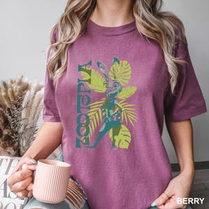 May include: Berry-colored t-shirt featuring a graphic of a rabbit character from Zootopia, surrounded by green tropical leaves. The word "Zootopia" is printed vertically in teal. The shirt is a comfortable, casual style.