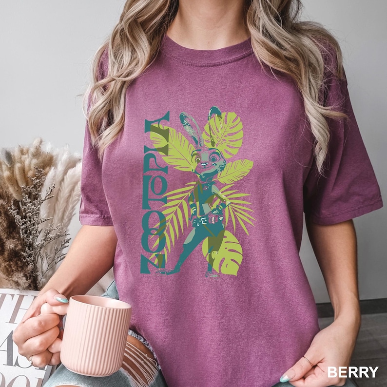 May include: Berry-colored t-shirt featuring a graphic of a rabbit character from Zootopia, surrounded by green tropical leaves. The word "Zootopia" is printed vertically in teal. The shirt is a comfortable, casual style.