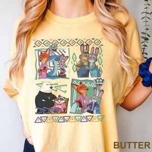 K&ouml;nnte beinhalten: Gelbes T-Shirt mit einem farbenfrohen Grafikdesign von Charakteren aus dem Animationsfilm Zootopia. Das Design zeigt verschiedene Tierfiguren in verschiedenen Szenen, mit geometrischen Mustern und dem Wort "BUTTER" unten rechts.