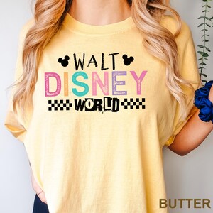 May include: A butter yellow short-sleeve t-shirt featuring the black text WALT DISNEY WORLD with two black Mickey Mouse ear silhouettes. The word DISNEY is printed in large, multi-colored letters including pink, orange, teal, and purple.