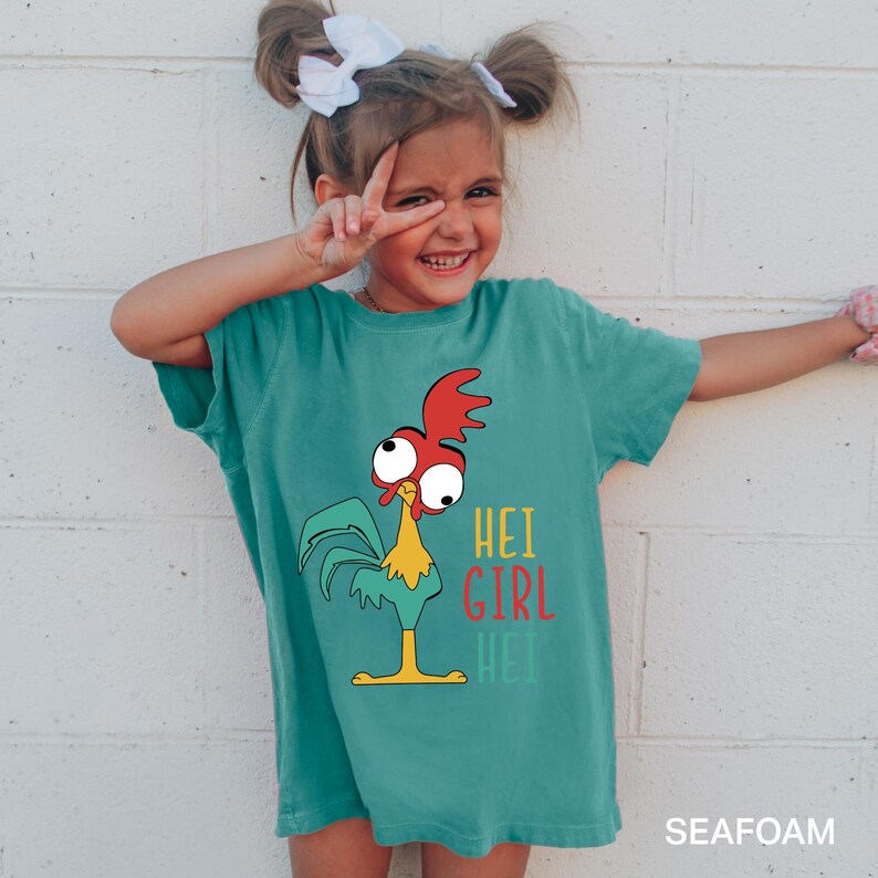 May include: Seafoam green t-shirt featuring a cartoon rooster with the text "HEI GIRL HEI". The rooster has a red comb, big eyes, and a yellow beak. The shirt is a comfortable fit.