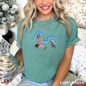 Op de afbeelding: Lichtgroen T-shirt met een geborduurd ontwerp met een vos en een konijn die elkaar achternazitten, verbonden door een blauwe slangachtige figuur. De tekst "LIGHT GREEN" staat onderaan het shirt.