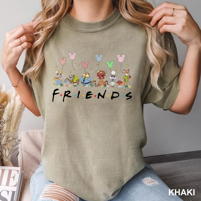 K&ouml;nnte beinhalten: Khakifarbenes T-Shirt mit dem Wort "FRIENDS" in einer stilisierten Schriftart. &Uuml;ber dem Wort befinden sich Cartoon-Tierfiguren, die Mickey Mouse-f&ouml;rmige Ballons halten. Das Shirt ist ein bequemer, l&auml;ssiger Stil.