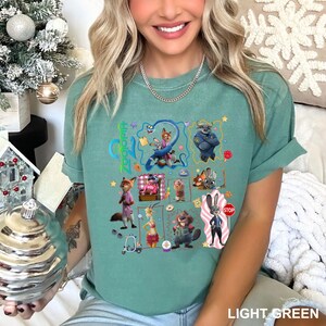 May include: A light green t-shirt featuring a graphic print of various animated characters from the movie Zootopia, including Nick Wilde and Judy Hopps, arranged in a collage style with small icons like a stop sign and a scooter.