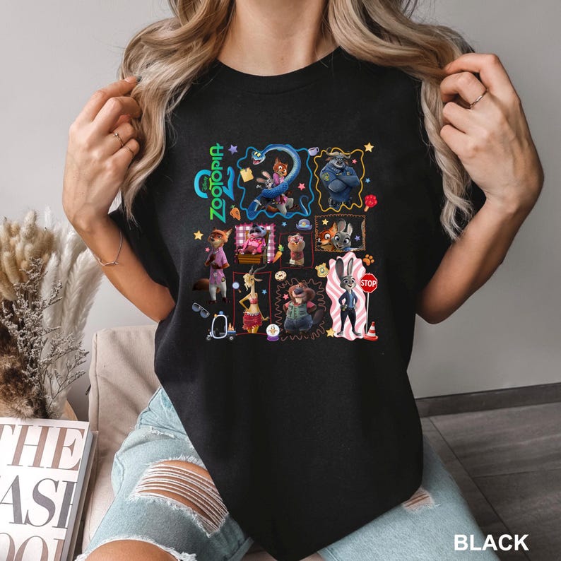 May include: A black t-shirt featuring a colorful graphic print of various characters from the movie Zootopia. The design includes multiple framed illustrations of animals, the Zootopia logo, and small icons like a stop sign, a donut, and a carrot.