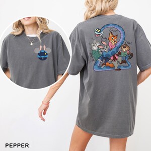 May include: Pepper-colored t-shirt featuring a cartoon design. The front has a blue and yellow cartoon character. The back has a blue snake with cartoon characters and names. The shirt is a comfortable, oversized fit.