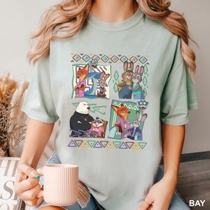 K&ouml;nnte beinhalten: Hellgr&uuml;nes T-Shirt mit einem farbenfrohen Grafikdesign mit Charakteren aus dem Animationsfilm Zoomania. Das Design enth&auml;lt Bilder eines Fuchses, eines Kaninchens, eines B&auml;ren und anderer Charaktere in einem quadratischen Muster. Das Wort "BAY" ist unten rechts aufgedruckt.