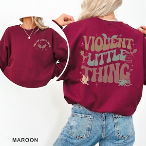 May include: Maroon sweatshirt with the text "VIOLENT LITTLE THING" on the back, along with dragon and book graphics. The front of the sweatshirt has the same text and dragon graphic. The sweatshirt is a solid color.