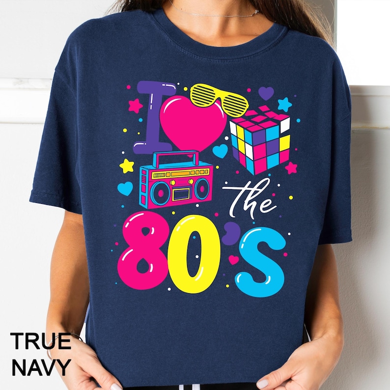 May include: A true navy t-shirt featuring a colorful graphic print that reads I Love The 80s. The design includes a pink heart, yellow shutter shades, a multi-colored cube puzzle, a pink boombox, and various small hearts and stars in bright neon colors.