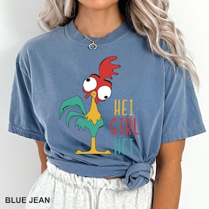 May include: Blue jean-colored t-shirt featuring a cartoon rooster with red, yellow, and green accents. The shirt has the text "HEI GIRL HEI" in a colorful font. The shirt is a comfortable, casual style.