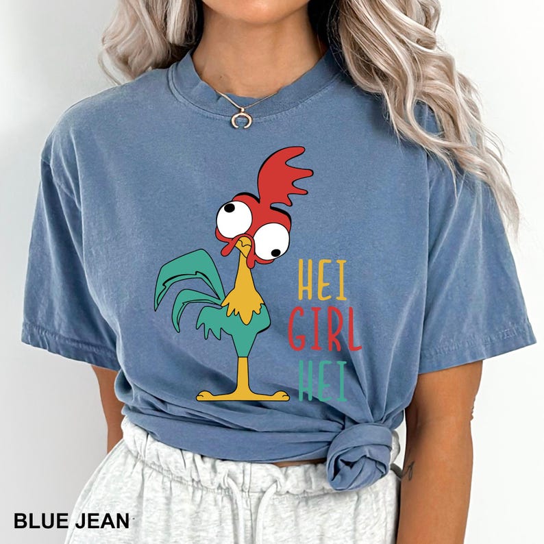 May include: Blue jean-colored t-shirt featuring a cartoon rooster with red, yellow, and green accents. The shirt has the text "HEI GIRL HEI" in a colorful font. The shirt is a comfortable, casual style.