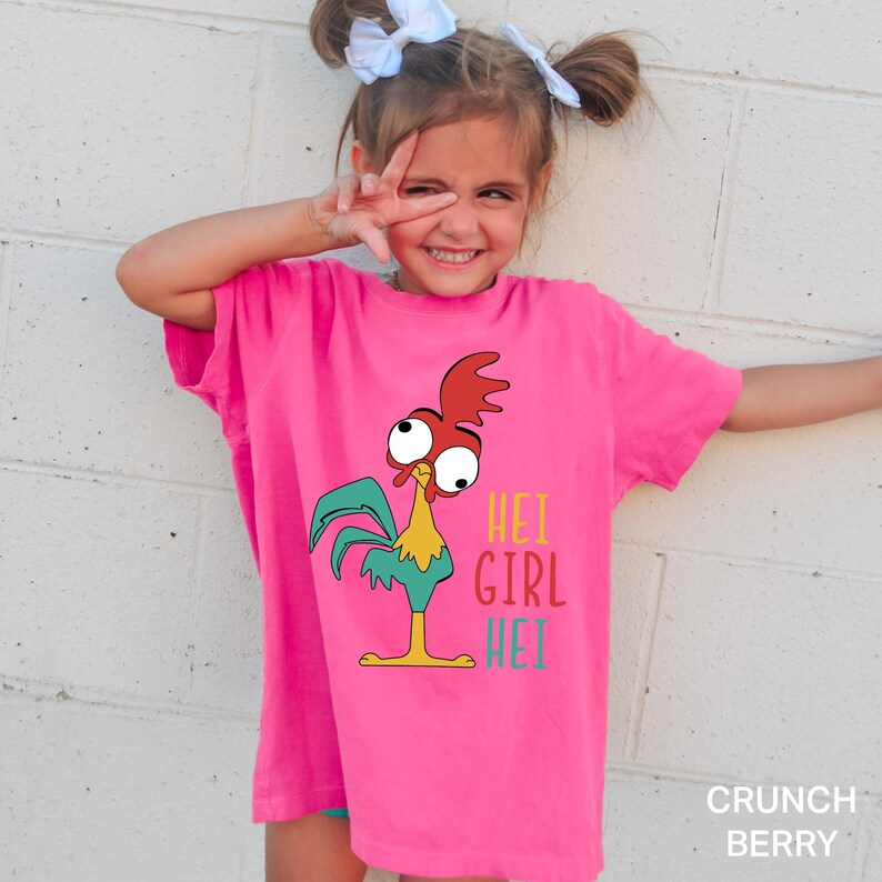 May include: A bright pink t-shirt featuring a cartoon rooster with the text "HEI GIRL HEI" in colorful letters. The rooster has red, yellow, and teal accents. The shirt is a comfortable fit.