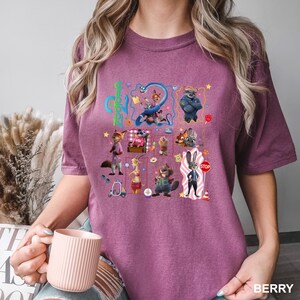 May include: A berry-colored short-sleeve t-shirt featuring a graphic print of various Zootopia characters, including Judy Hopps and Nick Wilde, arranged in a collage with the text Zootopia in bright green letters and a red stop sign.