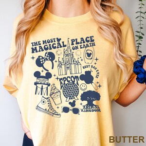 Puede incluir: Camiseta de color mantequilla con un dise&ntilde;o gr&aacute;fico azul marino. El dise&ntilde;o incluye el texto "The Most Magical Place on Earth" e iconos de Disney como un castillo y orejas de Mickey Mouse.