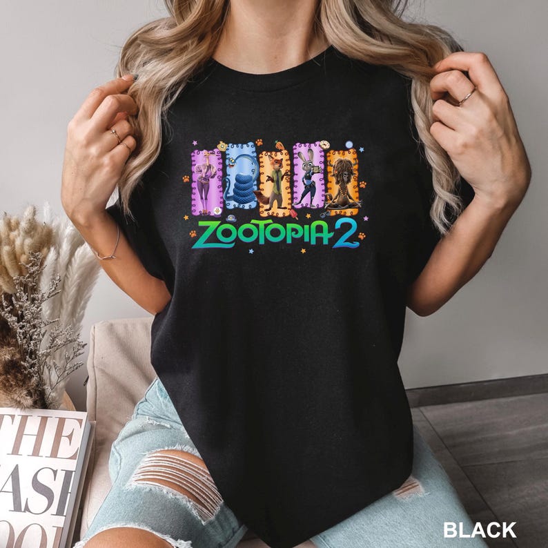 K&ouml;nnte beinhalten: Schwarzes T-Shirt mit einer farbenfrohen Grafik aus Zootopia 2. Das Design zeigt Charaktere in rechteckigen Rahmen &uuml;ber dem Text "ZOOTOPIA 2" in einer lebendigen, mehrfarbigen Schriftart. Das Wort "BLACK" ist unten aufgedruckt.