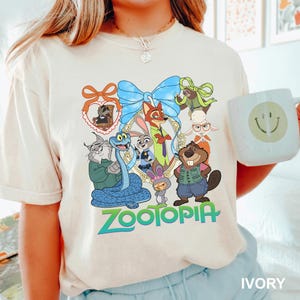 K&ouml;nnte beinhalten: Elfenbeinfarbenes T-Shirt mit einer farbenfrohen Grafik von Charakteren aus dem Animationsfilm "Zootopia". Das Design umfasst eine blaue Schlange, einen Fuchs, ein Kaninchen und andere Tierfiguren. Das Wort "Zootopia" ist in Gr&uuml;n und Blau gedruckt.