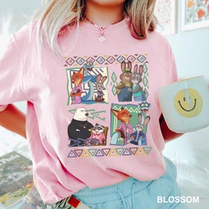 K&ouml;nnte beinhalten: Hellrosa T-Shirt mit einem farbenfrohen Grafikdesign von Zeichentrickfiguren. Das Design ist in mehrere Felder unterteilt, die verschiedene Charaktere und geometrische Muster zeigen. Eine wei&szlig;e Tasse mit einem Smiley wird im Bild gehalten.