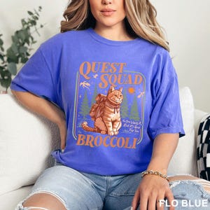 May include: A blue t-shirt with a graphic featuring a cat wearing a backpack, the words "QUEST SQUAD" and "BROCCOLI". The graphic also includes the text "No One Wants It, But It's Good For You".