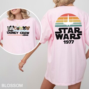 May include: Pale pink t-shirt with a Disney Crew graphic on the front and a Star Wars design on the back. The Disney Crew graphic features characters and the text "DISNEY CREW". The Star Wars design includes a sunset, a spaceship, and the text "STAR WARS 1977".