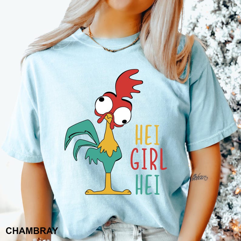 May include: Light blue t-shirt featuring a cartoon rooster with large eyes, red comb, and teal feathers. The text "HEI GIRL HEI" is printed in colorful letters. The shirt is made of a soft material.